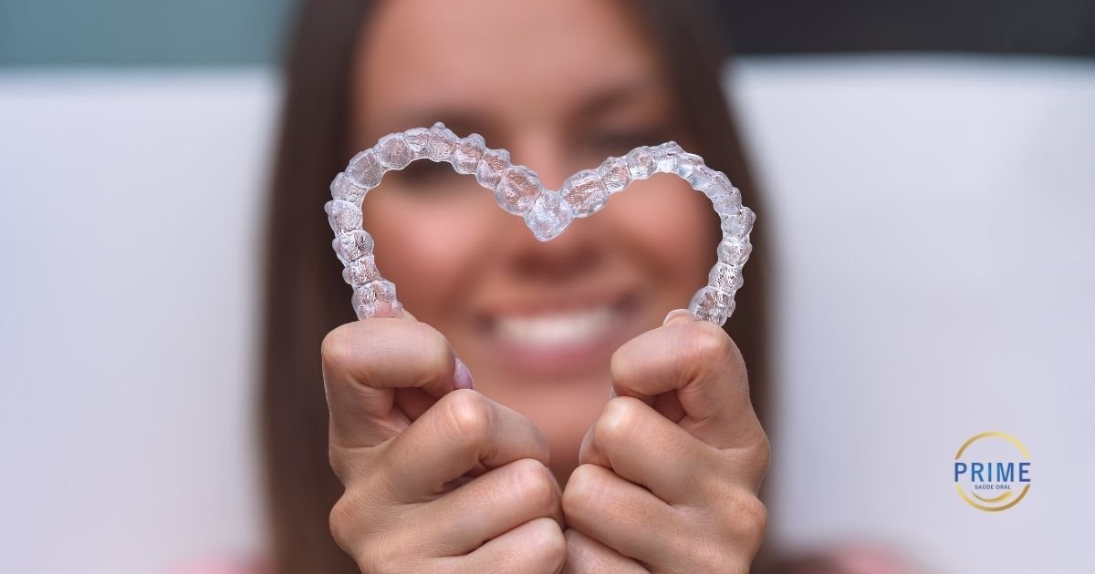 Mãos segurando um alinhador Invisalign no formato de coração com uma mulher sorrindo ao fundo, simbolizando o cuidado e o planejamento digital do tratamento.