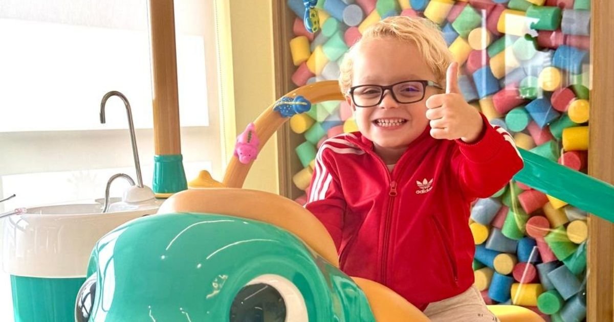 Menino loiro de óculos e casaco vermelho sentado na cadeira odontológica customizada em formato de dinossauro verde, sorrindo e fazendo sinal de "joinha".