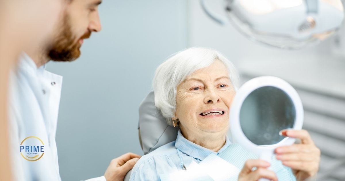Paciente idosa de cabelos brancos sorrindo no consultório odontológico enquanto segura um espelho e recebe atenção do dentista, com logo da Prime Saúde Oral