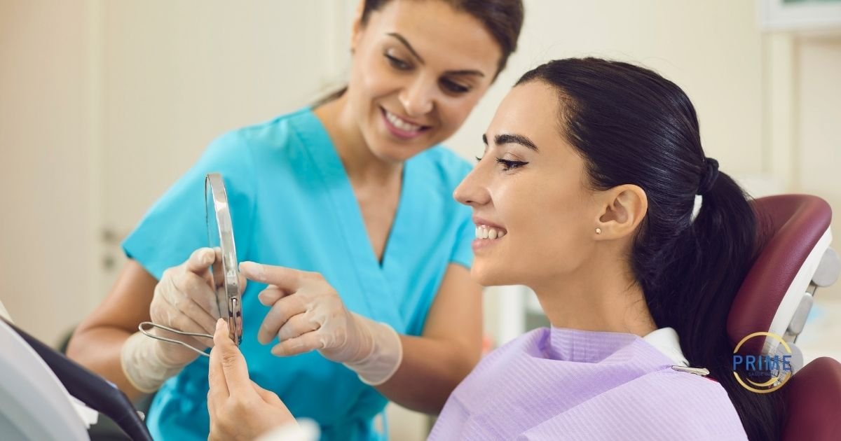 Dentista de uniforme azul mostrando o resultado do tratamento no espelho para paciente jovem sorridente na cadeira odontológica, com logo da Prime Saúde Oral