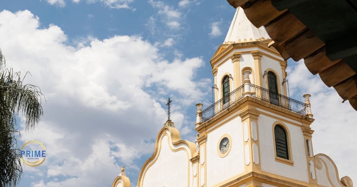 Fachada da igreja histórica colonial de Santana de Parnaíba com torre, relógio e céu azul ao fundo, com logo da Prime Saúde Oral