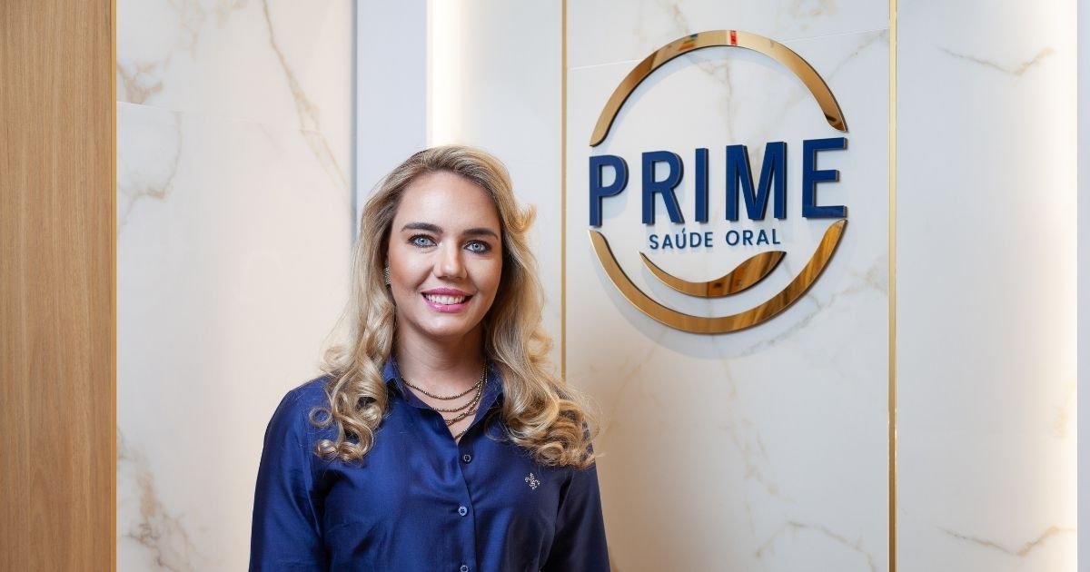 Dra. Vanessa Nani sorrindo em frente à logo dourada da Prime Saúde Oral em parede de mármore branco na clínica em Alphaville
