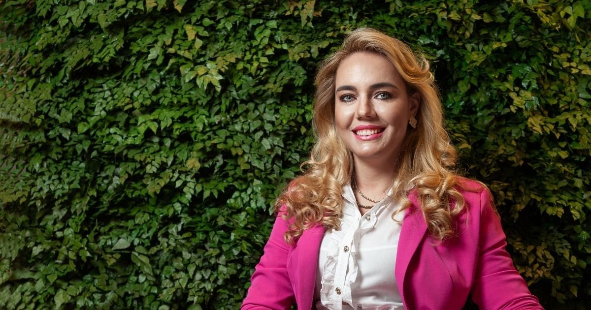 Dra. Vanessa Nani sorrindo em frente a parede coberta por vegetação verde, com blazer pink e blusa branca, com logo da Prime Saúde Oral