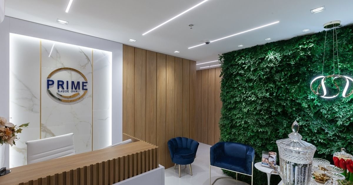 Recepção da Prime Saúde Oral em Alphaville com logo dourada na parede de mármore, painel de madeira, jardim vertical e poltronas azuis em ambiente moderno e sofisticado