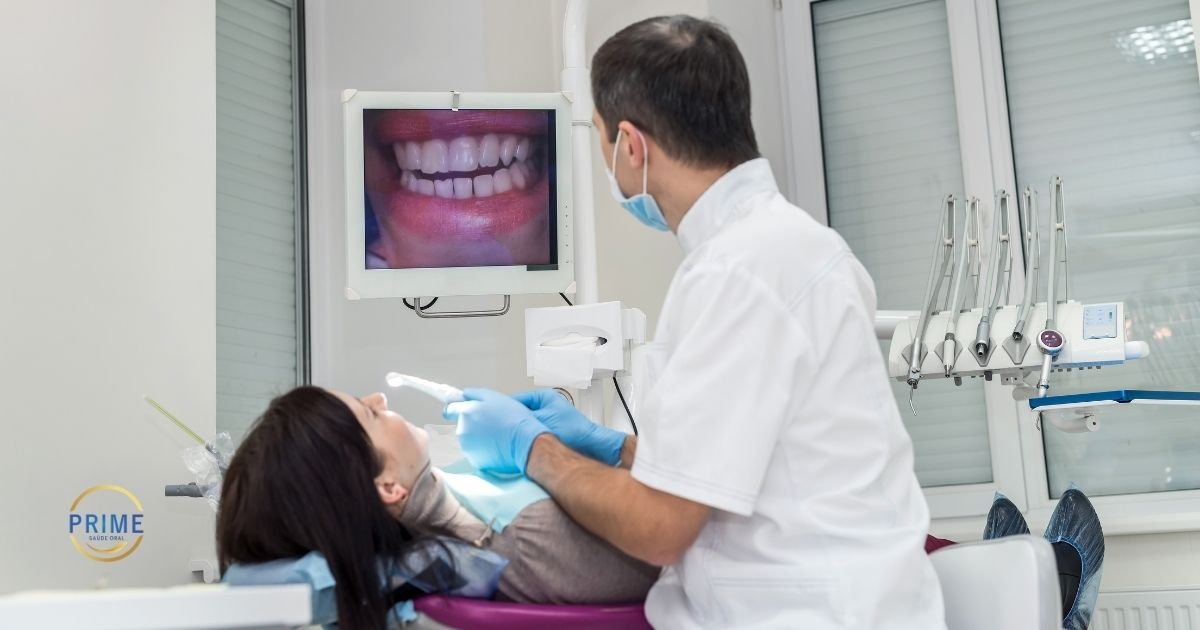 Dentista com máscara e luvas azuis utilizando câmera intraoral em paciente enquanto monitor exibe imagem ampliada do sorriso em tempo real, com logo da Prime Saúde Oral