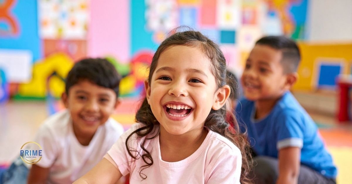 Grupo de crianças sorrindo em ambiente escolar colorido, com menina em destaque mostrando sorriso saudável com dentes de leite, com logo da Prime Saúde Oral