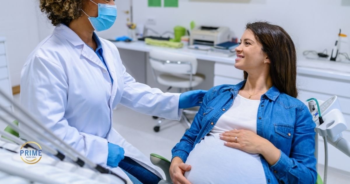 Dentista com jaleco branco e máscara conversando de forma acolhedora com gestante sorridente sentada na cadeira odontológica, com mão na barriga, com logo da Prime Saúde Oral