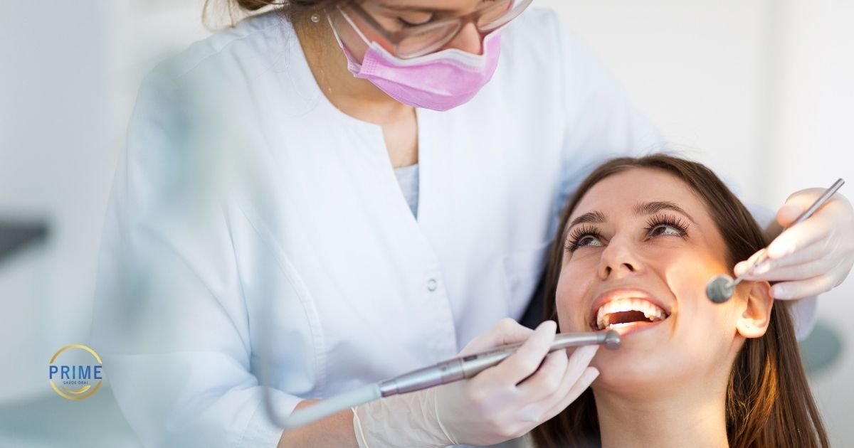 Dentista com máscara rosa e luvas brancas realizando limpeza dental em paciente jovem sorridente reclinada na cadeira odontológica, com logo da Prime Saúde Oral